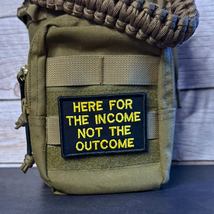 An olive green tactical pouch with a black and yellow embroidered patch reading 'HERE FOR THE INCOME NOT THE OUTCOME', shown with a paracord strap against a rustic wood background.