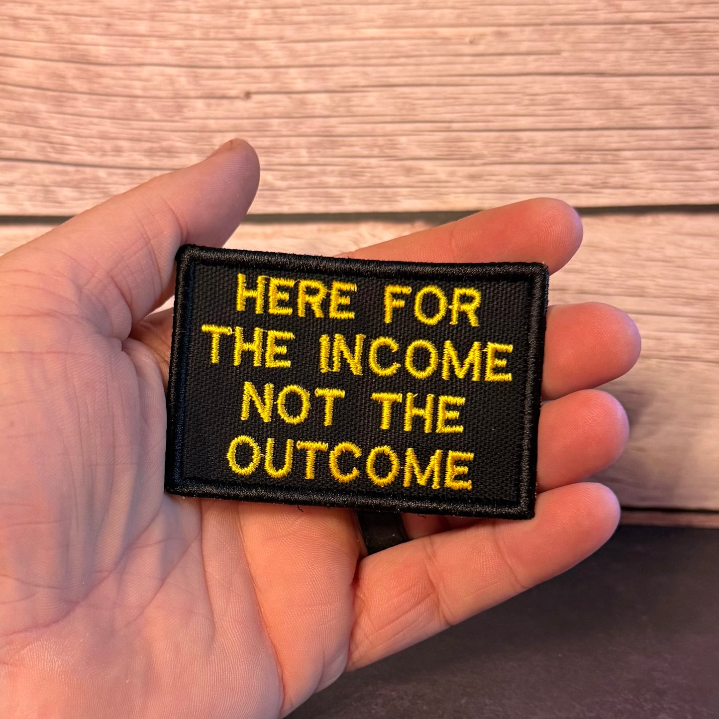 A hand holding a rectangular black embroidered patch with yellow text that reads 'HERE FOR THE INCOME NOT THE OUTCOME' against a wood plank background.