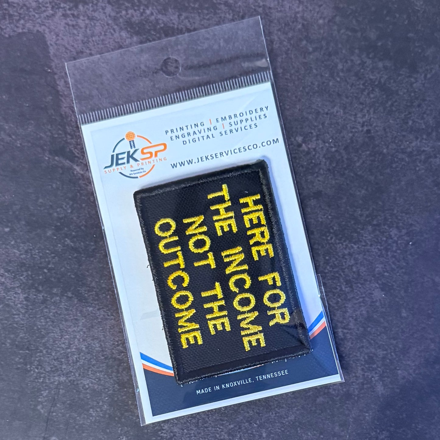 A black rectangular embroidered patch with yellow text reading 'HERE FOR THE INCOME NOT THE OUTCOME' in diagonal lettering, sealed in a clear plastic package with a white backing card branded JEKSP.