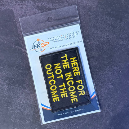 A black rectangular embroidered patch with yellow text reading 'HERE FOR THE INCOME NOT THE OUTCOME' in diagonal lettering, sealed in a clear plastic package with a white backing card branded JEKSP.