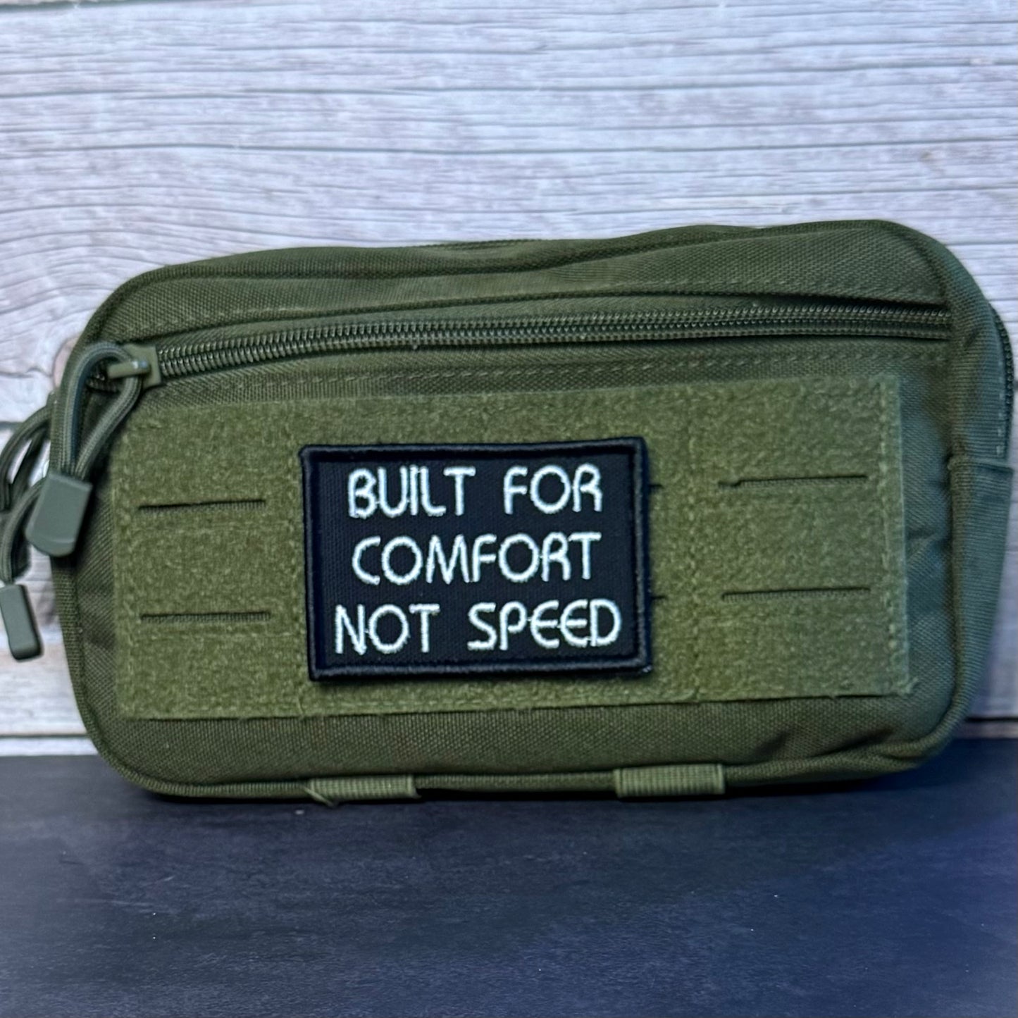 An olive green tactical pouch with a black rectangular patch that reads 'BUILT FOR COMFORT NOT SPEED' in white text, shown against a light gray wood background.