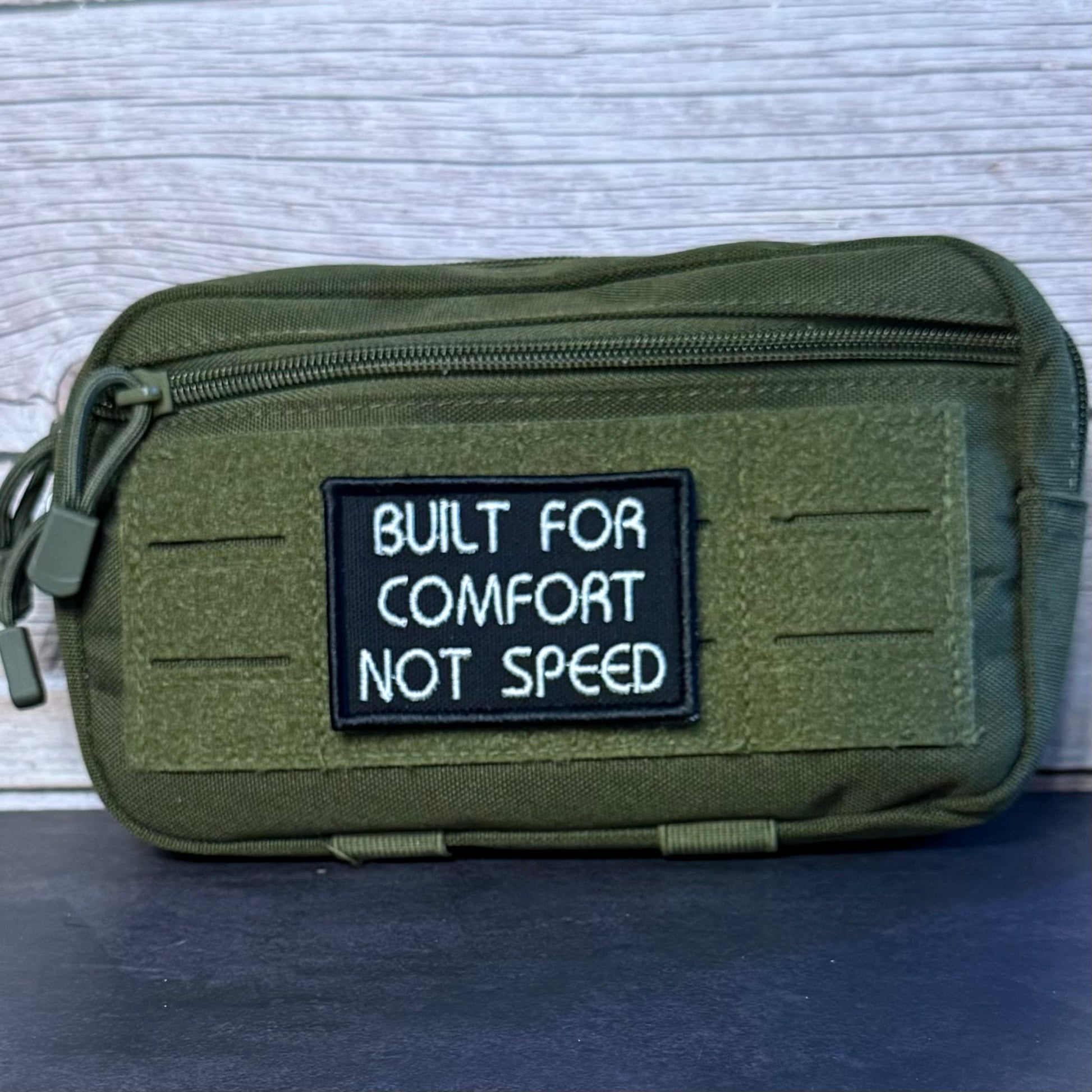An olive green tactical pouch with a black rectangular patch that reads 'BUILT FOR COMFORT NOT SPEED' in white text, shown against a light gray wood background.