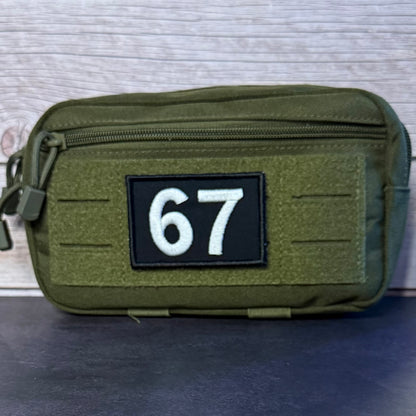 An olive green tactical pouch with a top zipper and a black rectangular patch featuring the white numbers '67', set against a light gray wood-textured background.