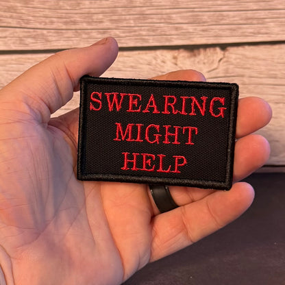 A hand holding a rectangular black embroidered patch with red text that reads 'SWEARING MIGHT HELP' against a wood-paneled background.