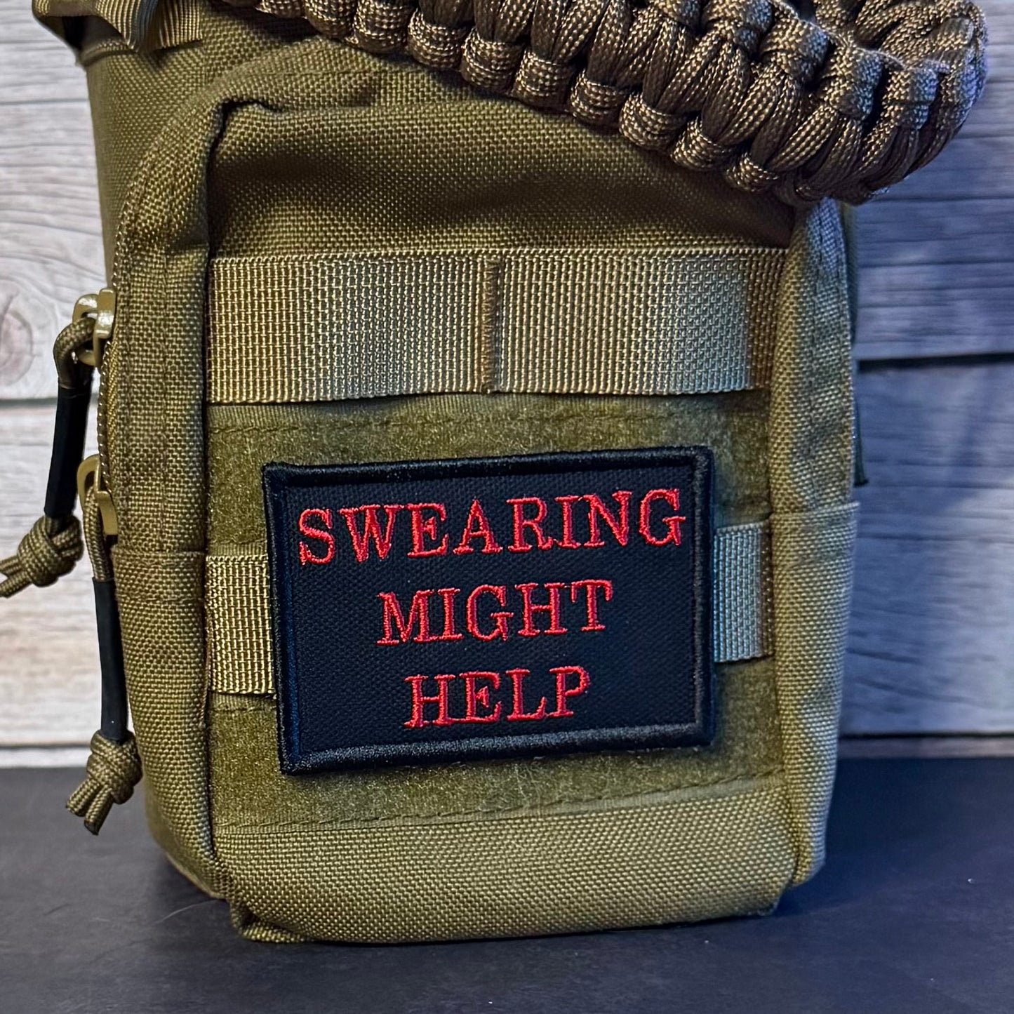 An olive green tactical pouch with a black rectangular patch featuring red embroidered text that reads 'SWEARING MIGHT HELP'. The pouch has a woven strap across the top and a zipper pull on the left side, photographed against a rustic wood background.