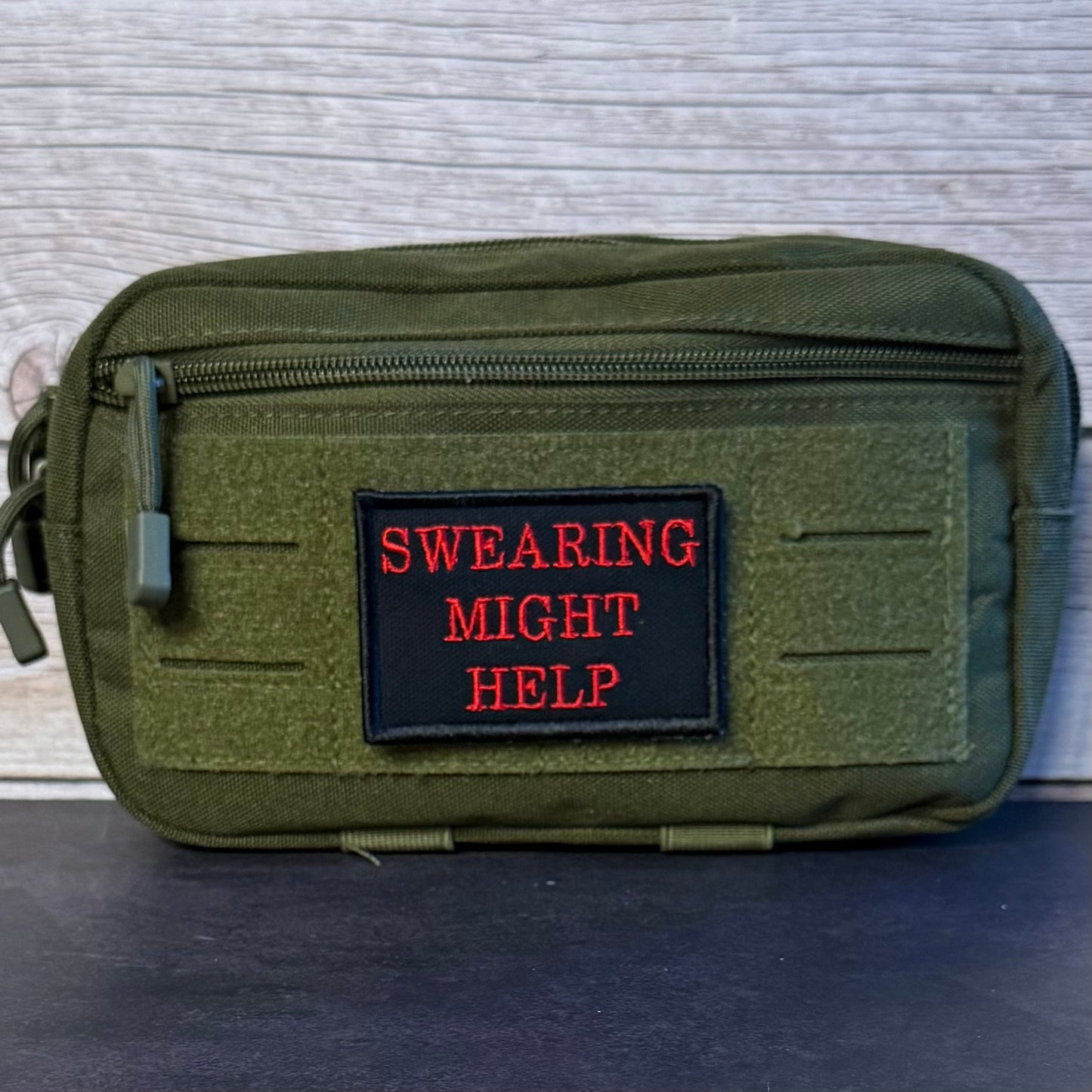 An olive green tactical pouch with a black rectangular patch featuring the red embroidered text 'SWEARING MIGHT HELP'. The pouch has a top zipper, MOLLE webbing, and a green zipper pull, photographed against a light gray wood panel background.