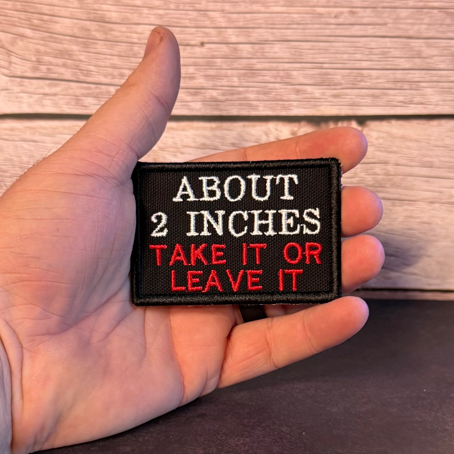A hand holding a rectangular black embroidered patch with the text 'ABOUT 2 INCHES TAKE IT OR LEAVE IT' in white and red lettering, set against a rustic wood plank background.