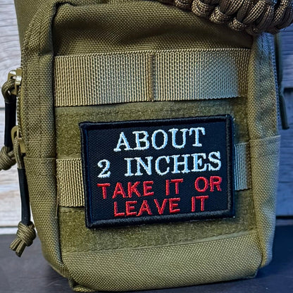 An olive green tactical pouch with a black embroidered patch that reads 'ABOUT 2 INCHES TAKE IT OR LEAVE IT' in white and red text. A thick brown paracord strap is wrapped around the top, set against a rustic gray wood plank background.