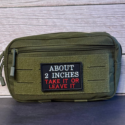 An olive green tactical pouch with a black rectangular patch on the front that reads 'ABOUT 2 INCHES TAKE IT OR LEAVE IT' in white and red text, photographed against a gray wood plank background.