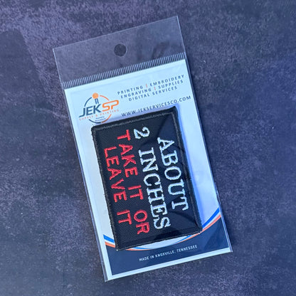 A black rectangular embroidered patch with red and white text reading 'ABOUT 2 INCHES TAKE IT OR LEAVE IT,' sealed in a clear plastic package with a white and blue backing card. The card shows the brand JEKSP.