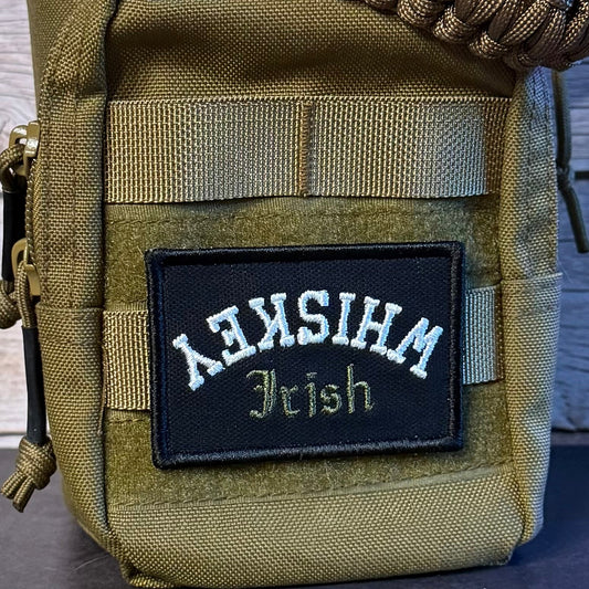 A close-up of a tactical utility pouch in olive drab green webbing, featuring a black embroidered patch with white and gold text reading 'WHISKEY Irish'. A thick braided tan strap is draped across the top, and the pouch is set against a rustic wooden background.