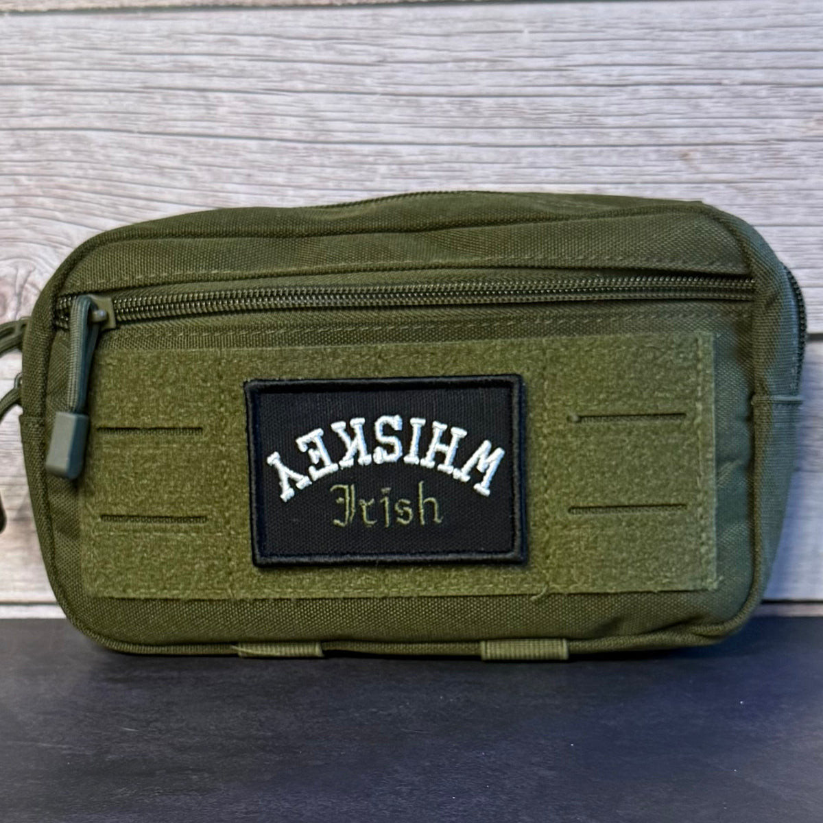 An olive green tactical pouch with a black rectangular patch reading 'Whiskey Irish' in white and gold lettering, set against a light wood background.