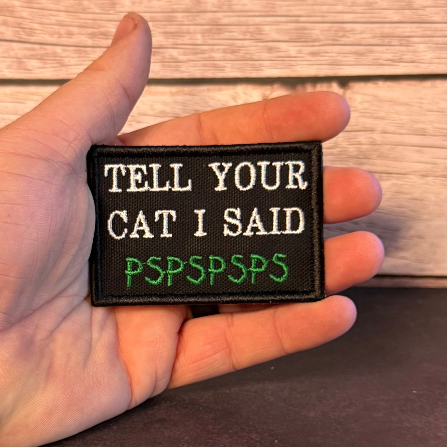 A hand holding a rectangular black embroidered patch with white text reading 'TELL YOUR CAT I SAID' and green text reading 'PSPSPSPS' against a wood plank background.