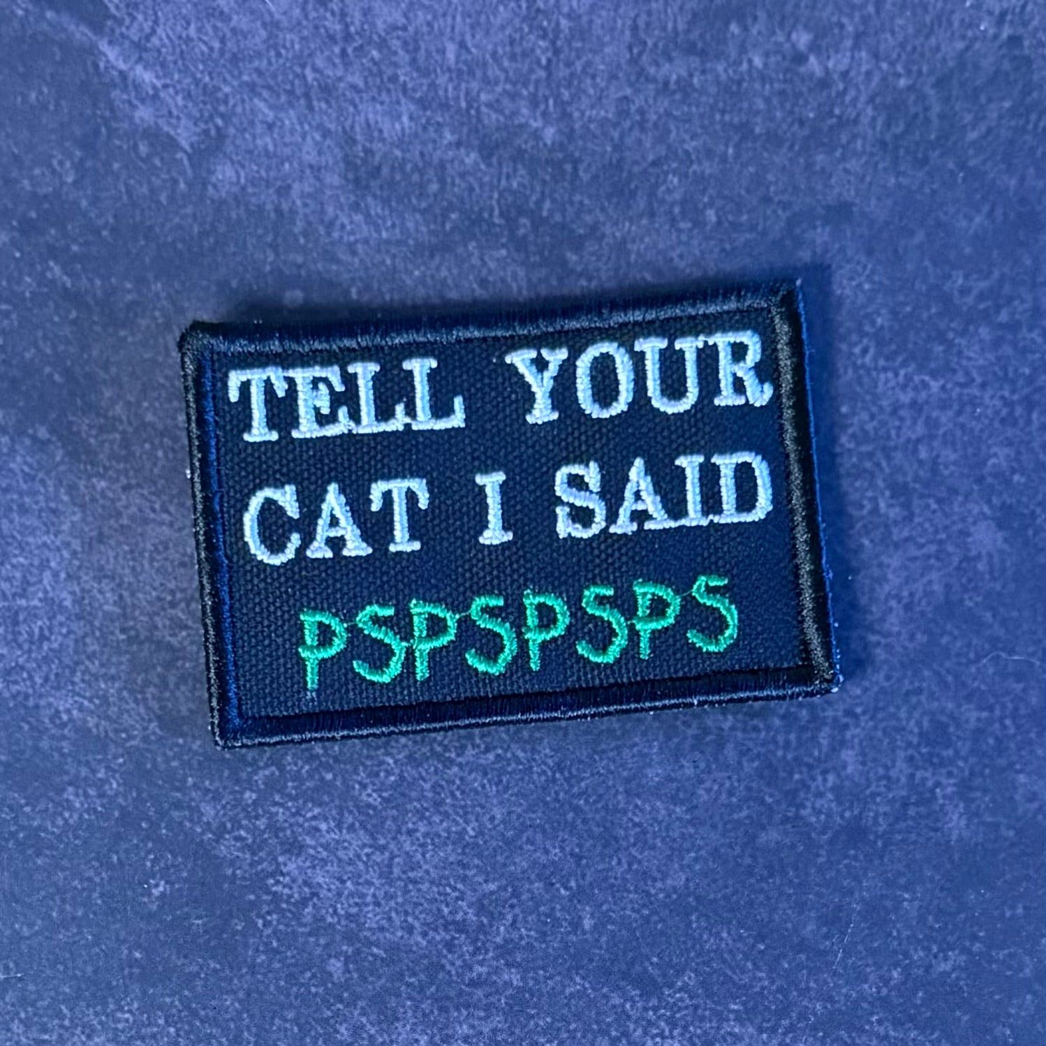 A rectangular embroidered patch with the text 'TELL YOUR CAT I SAID PSPSPSPS' in white and green lettering on a black background, placed on a textured blue surface.