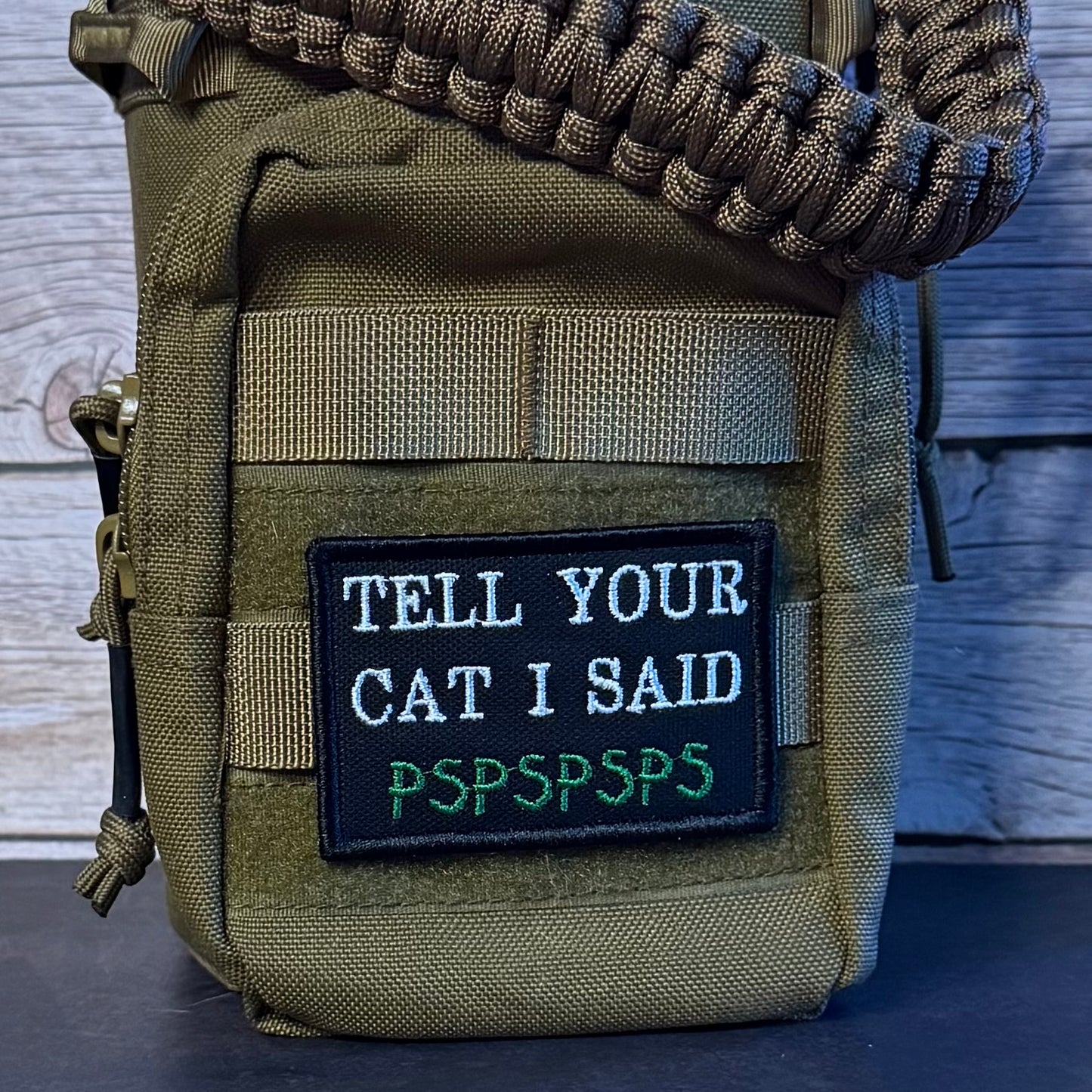 An olive green tactical pouch with a black embroidered patch reading 'TELL YOUR CAT I SAID PSPSPSPS' in white and green, featuring a paracord strap across the top, set against a rustic wood background.