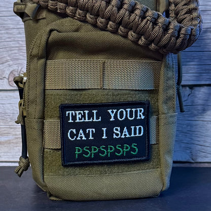 An olive green tactical pouch with a black embroidered patch reading 'TELL YOUR CAT I SAID PSPSPSPS' in white and green, featuring a paracord strap across the top, set against a rustic wood background.