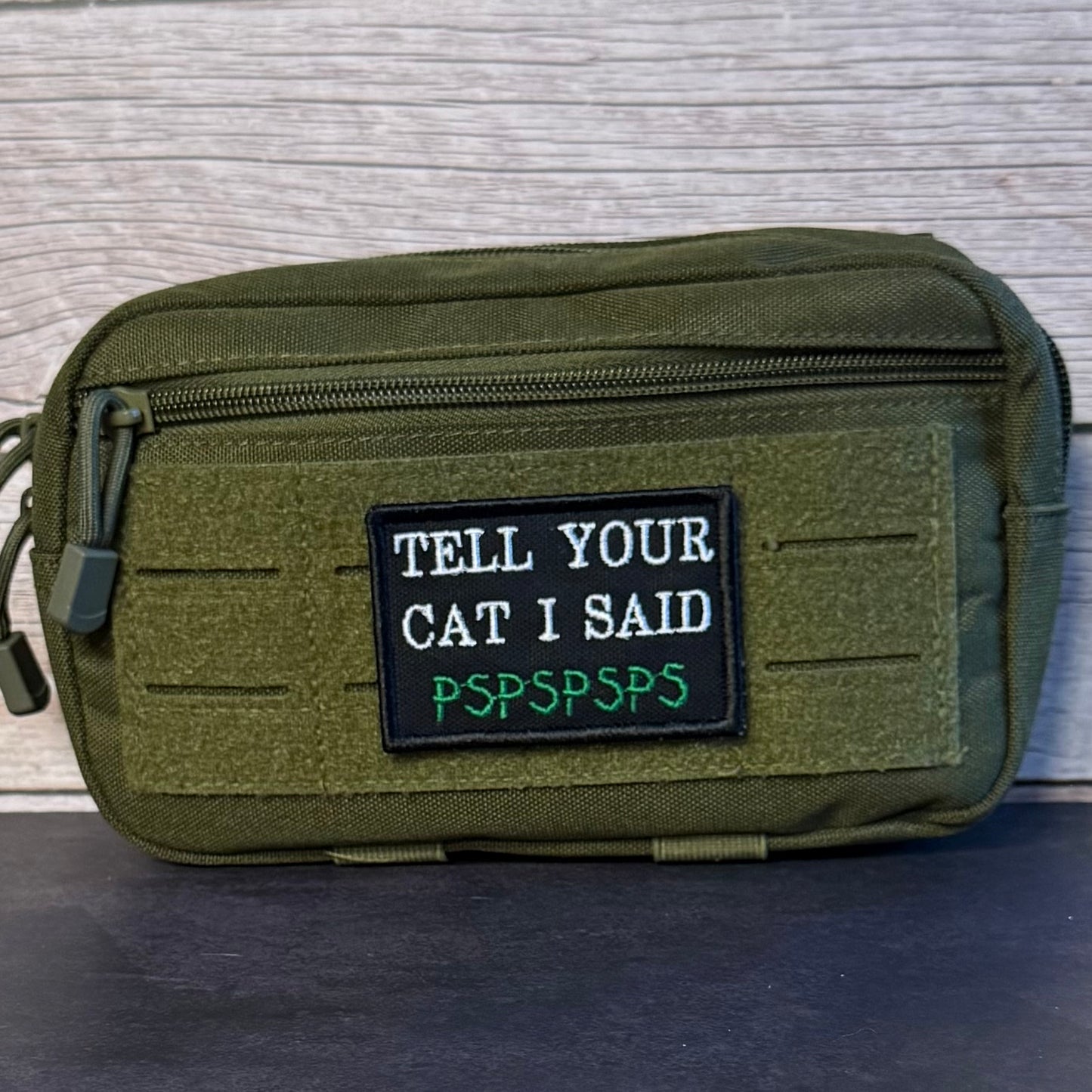 An olive green tactical pouch with a front zipper and a black rectangular patch that reads 'TELL YOUR CAT I SAID PSPSPSPS' in white and green text, set against a light wood background.