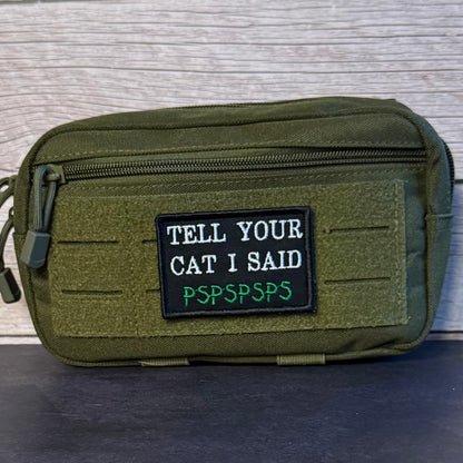 An olive green tactical pouch with a front zipper and a black rectangular patch that reads 'TELL YOUR CAT I SAID PSPSPSPS' in white and green text, set against a light wood background.