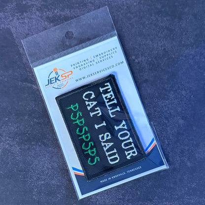 A black rectangular embroidered patch with white and green text reading 'TELL YOUR CAT I SAID JERSEYSPOONS' attached to a clear plastic retail card. The card shows the JEKSP brand and printed website text, and the package is photographed on a dark textured surface.