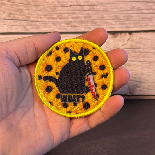 A circular embroidered patch held in a hand, featuring a black cat with yellow eyes holding a red knife, set against a yellow sunflower pattern with black spots and the word 'WHAT?' below.