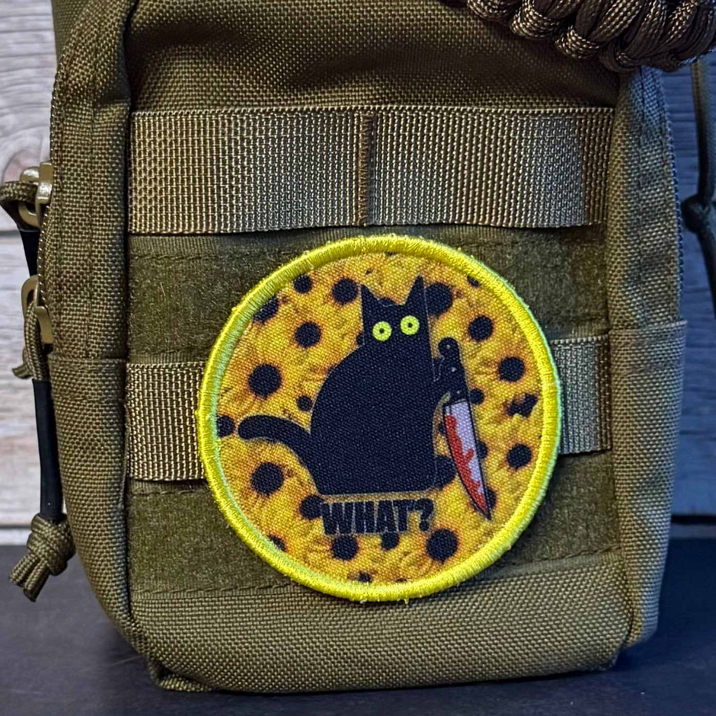 An olive drab tactical pouch with a paracord strap, featuring a circular embroidered patch of a black cat holding a knife on a yellow spotted background with the word 'WHAT?' below it.