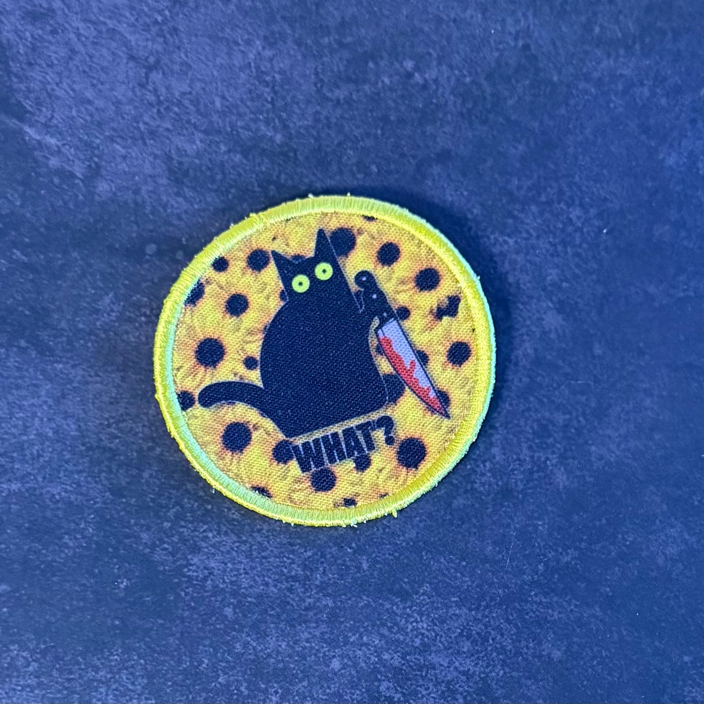 A circular embroidered patch featuring a black cat holding a knife, with the word 'WHAT?' below it, on a yellow background with black spots, set against a dark blue textured backdrop.