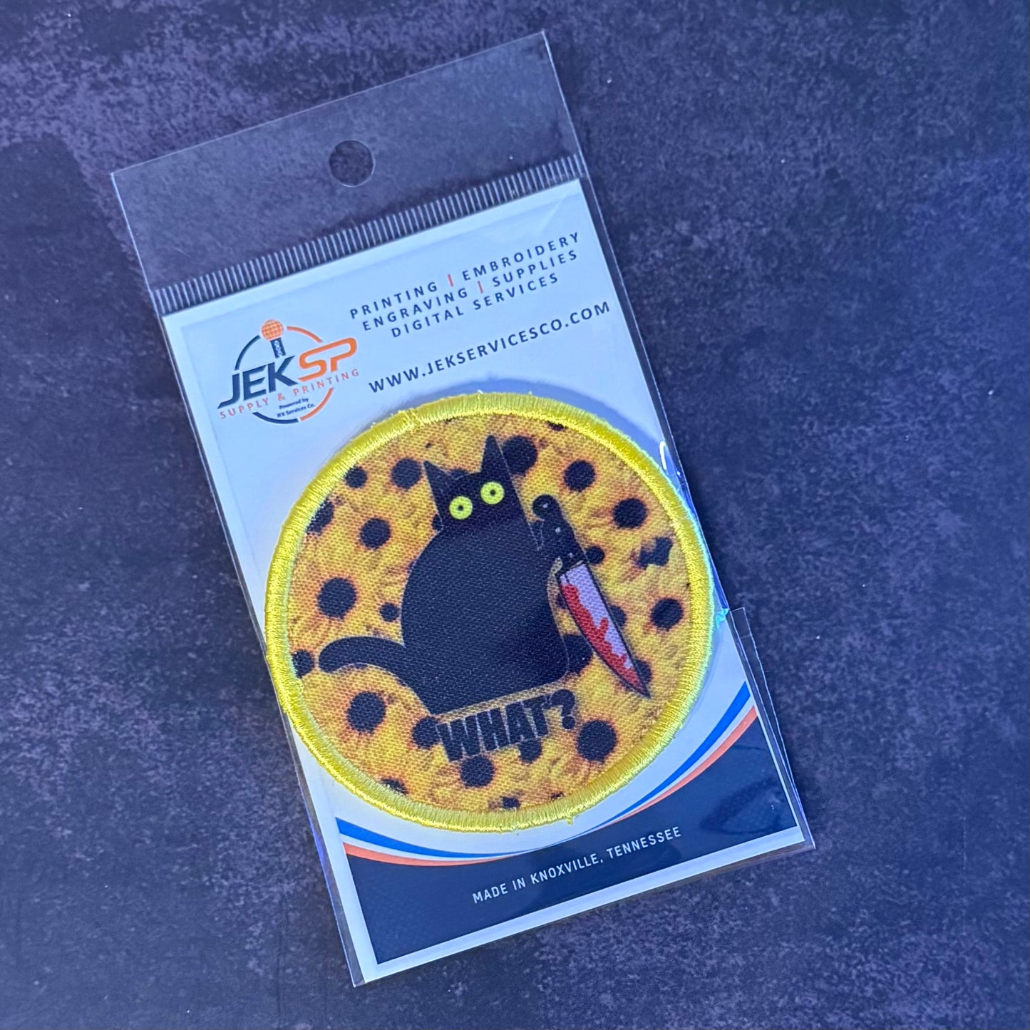 An embroidered circular patch featuring a black cat on a yellow spotted background with the word 'WHAT?' and a red chili pepper, shown in its clear retail packaging on a dark textured surface.