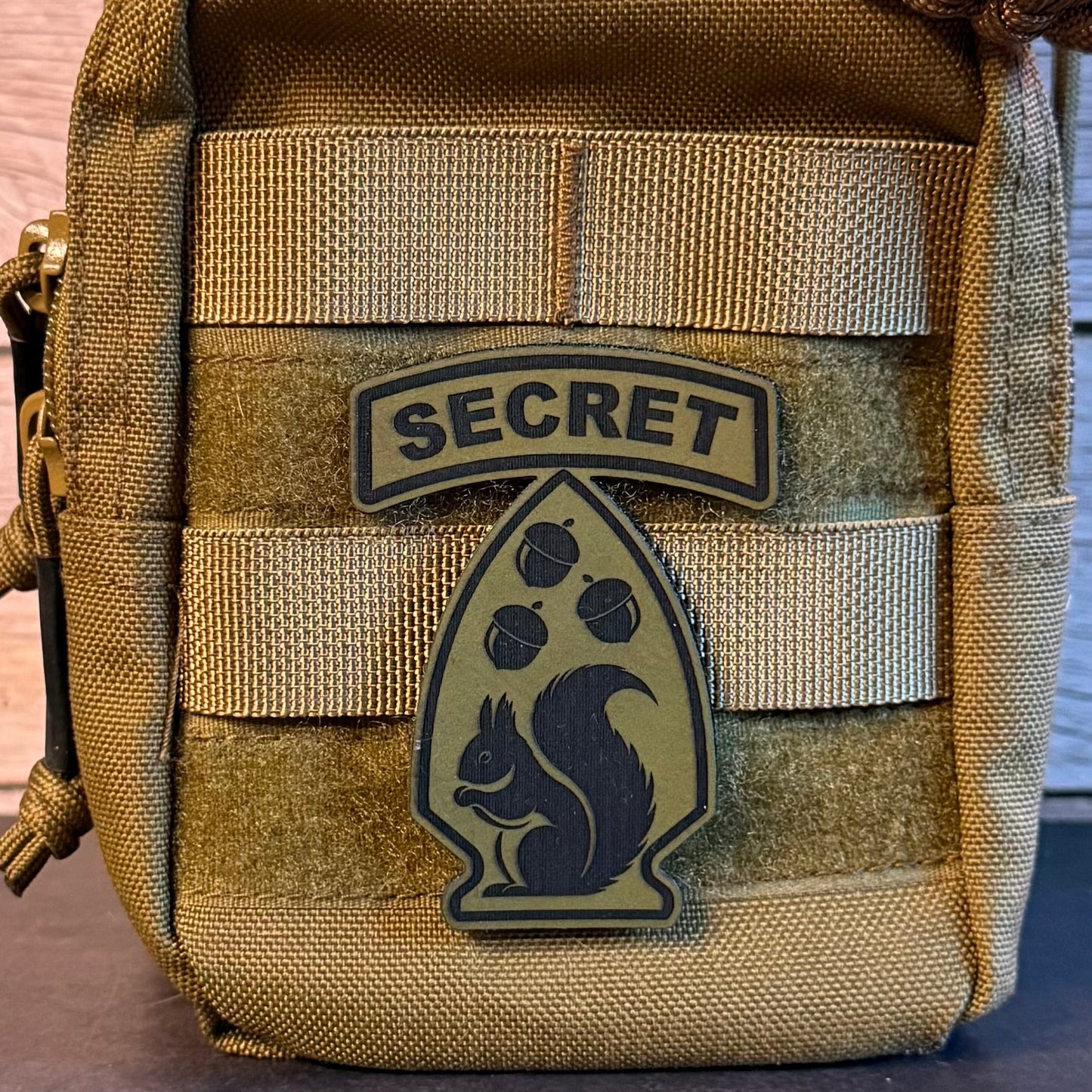 A close-up of a tan tactical pouch with MOLLE webbing, featuring a velcro patch that reads 'SECRET' above a squirrel emblem.