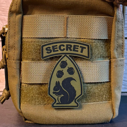 A close-up of a tan tactical pouch with MOLLE webbing, featuring a velcro patch that reads 'SECRET' above a squirrel emblem.