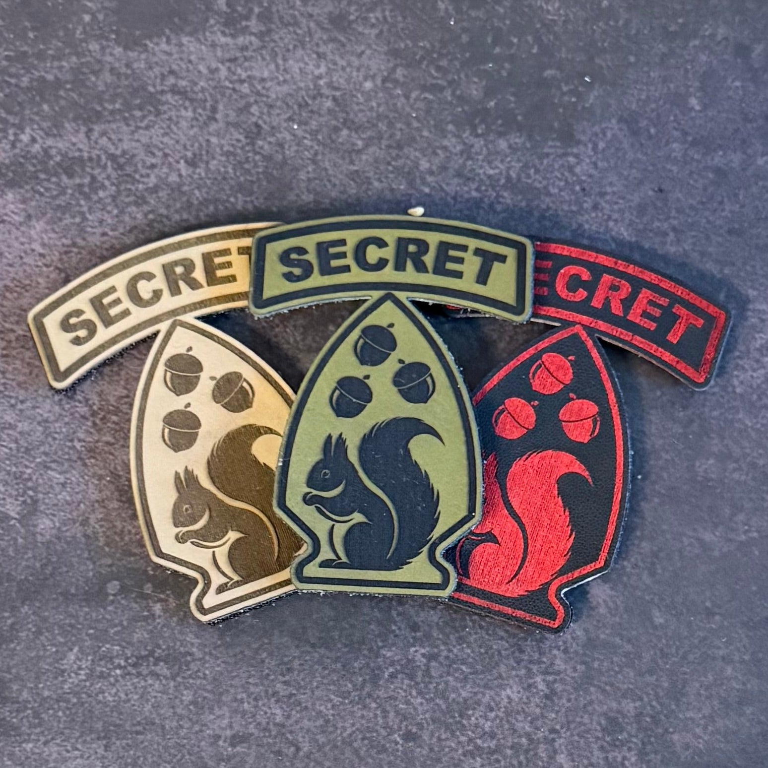 Three embroidered military-style patches with the word 'SECRET' and squirrel motifs, arranged on a dark textured surface.