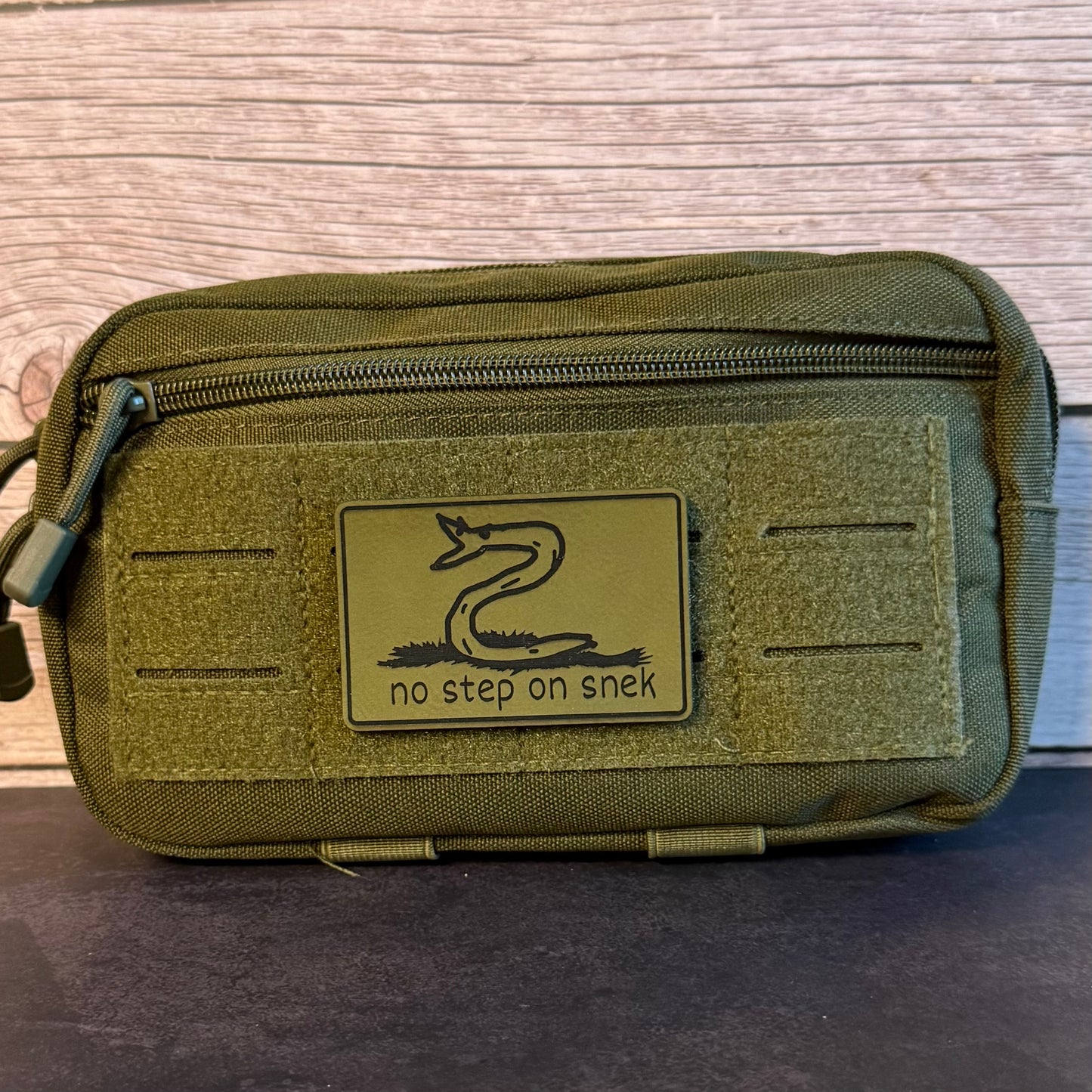 An olive green tactical pouch with a front zipper and a rectangular patch featuring a snake graphic and the text 'no step on snek'.