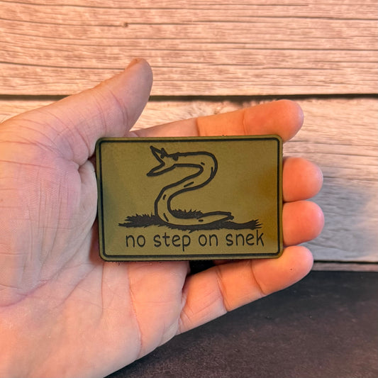 A hand holding a rectangular olive green rubber patch featuring a coiled snake and the text 'no step on snek' against a rustic wood plank background.