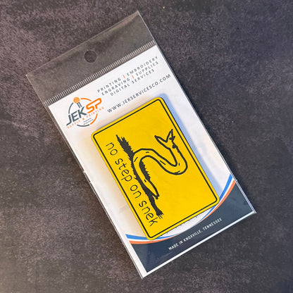 A yellow rectangular warning sticker with a black snake and the text 'no step on snax' is shown inside a clear plastic retail sleeve on a dark textured background. The sleeve includes branding for JER SERVICE.