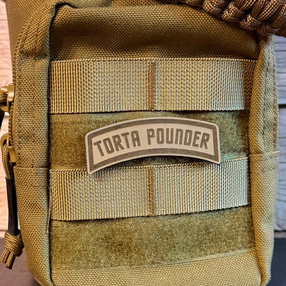 A close-up of a tactical pouch in olive drab and tan webbing, featuring a central patch that reads 'TORTA POUNDER' and a braided strap at the top, set against a rustic wood background.