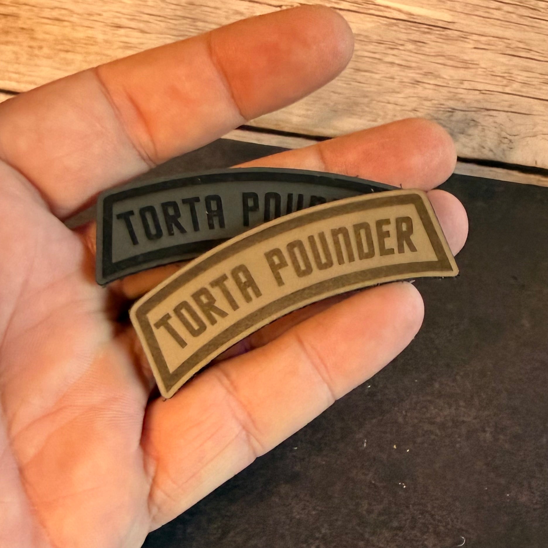Two curved PVC patch badges with the text 'TORTA POUNDER' held in a hand. One patch is dark gray/black and the other is tan/khaki, set against a background of wood and a dark surface.
