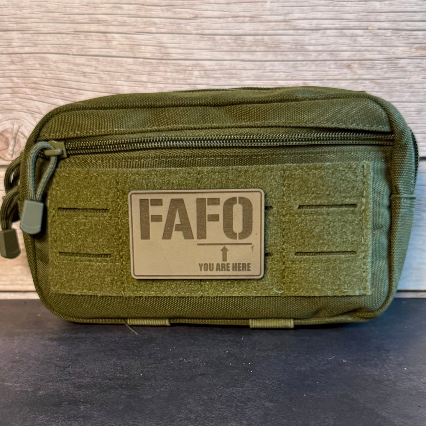 An olive green tactical pouch with a front zipper and a tan rectangular patch reading 'FAFO' and 'YOU ARE HERE', photographed against a wood plank background.