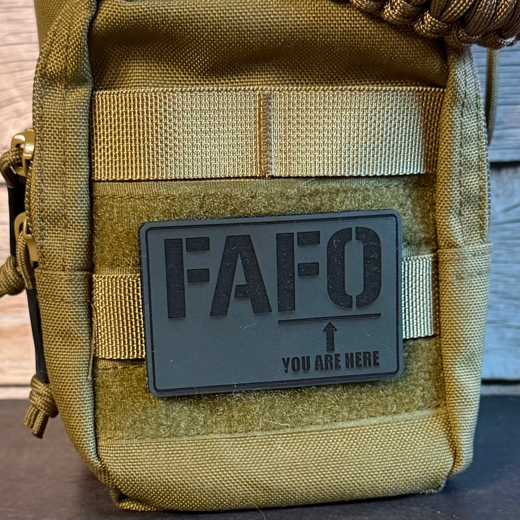 A close-up of a tactical pouch in olive drab and coyote tan with a dark gray rectangular patch that reads 'FAFO YOU ARE HERE' and an arrow, shown against a rustic wood background.