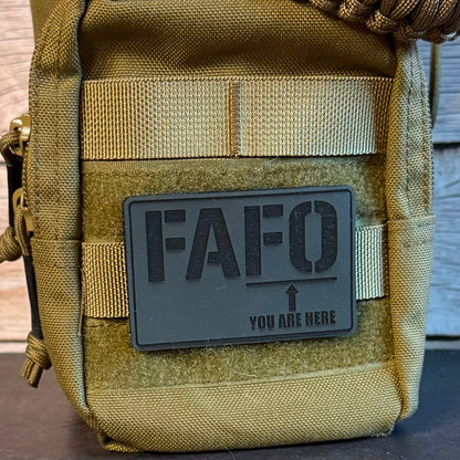A close-up of a tactical pouch in olive drab and coyote tan with a dark gray rectangular patch that reads 'FAFO YOU ARE HERE' and an arrow, shown against a rustic wood background.