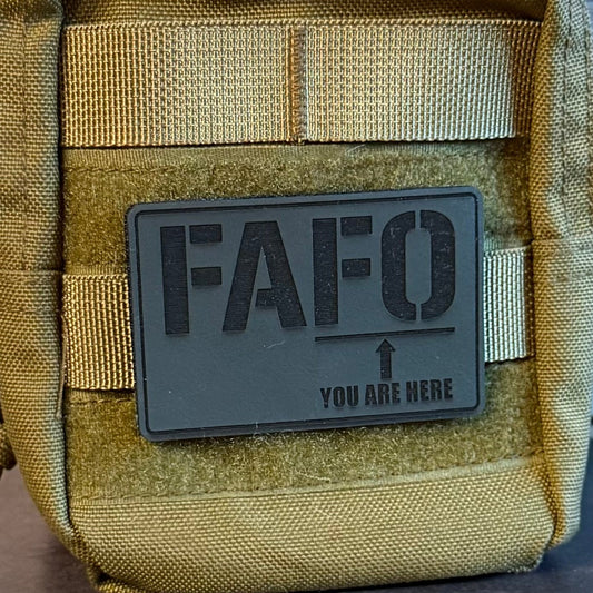 A close-up of a tactical pouch in olive drab and coyote tan with a dark gray rectangular patch that reads 'FAFO YOU ARE HERE' and an arrow, shown against a rustic wood background.