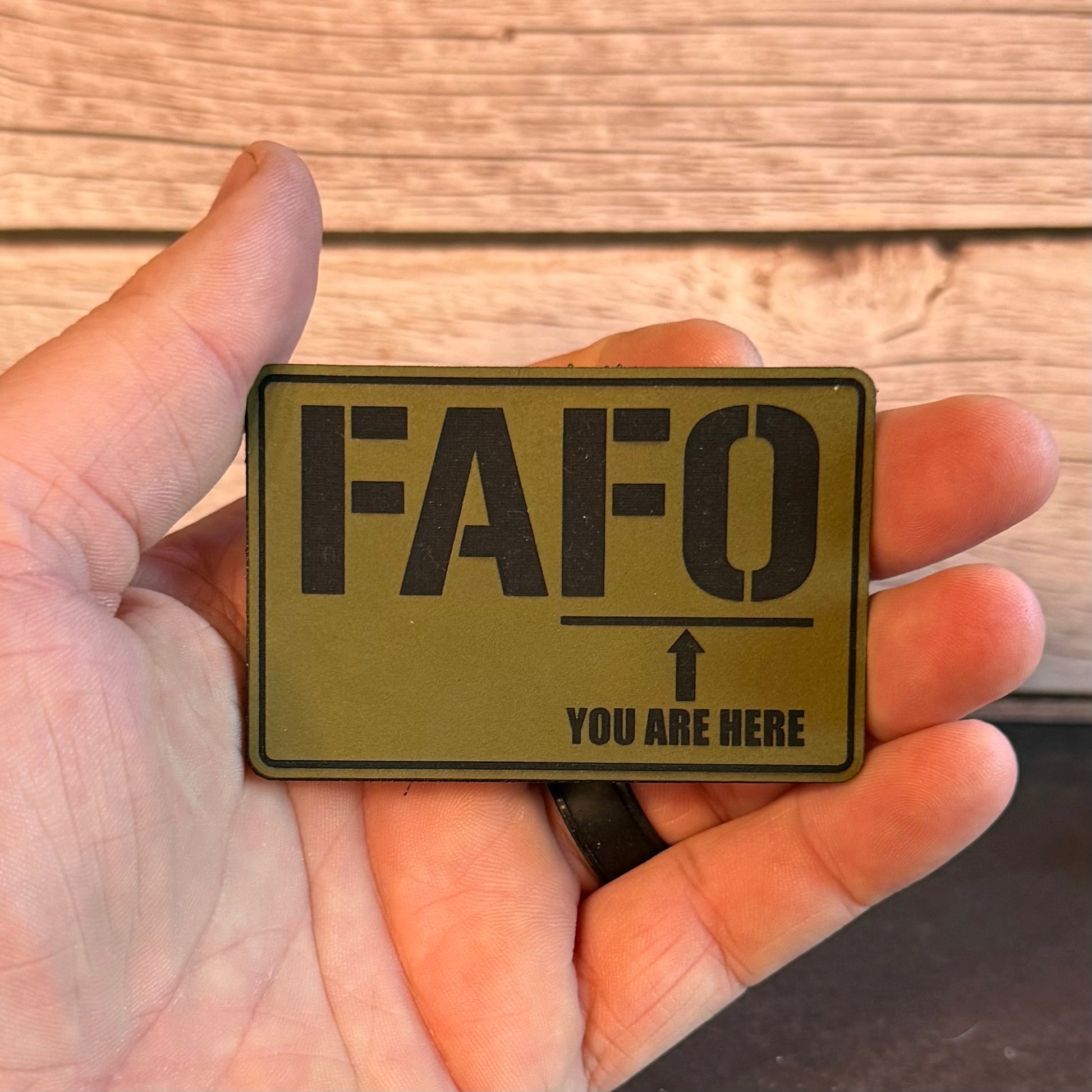 A hand holding a rectangular olive green tactical patch with black text reading 'FAFO' and 'YOU ARE HERE' with an arrow, against a wood plank background.