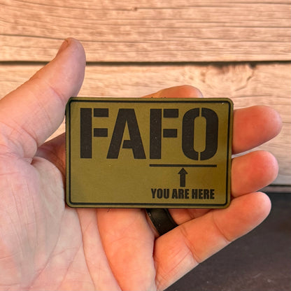 A hand holding a rectangular olive green tactical patch with black text reading 'FAFO' and 'YOU ARE HERE' with an arrow, against a wood plank background.