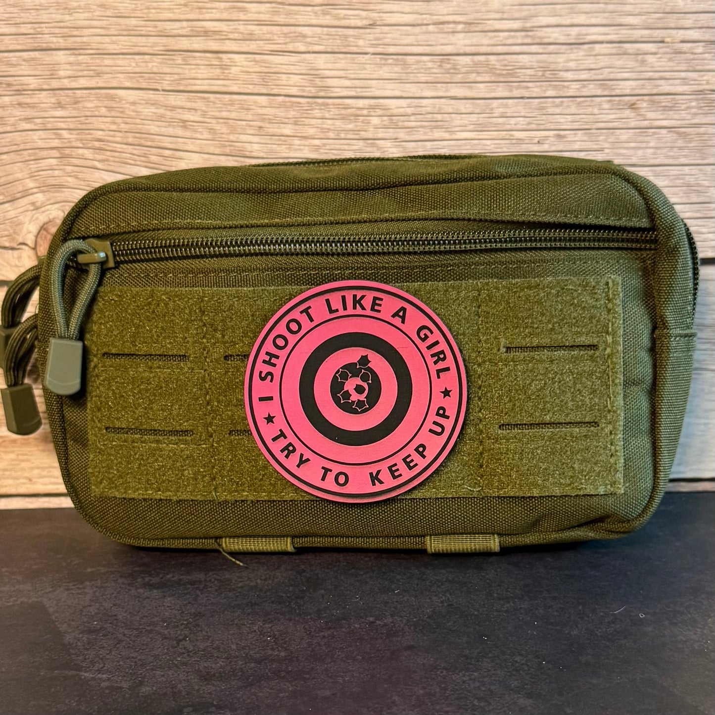 An olive green tactical pouch with a pink circular patch that reads 'I SHOOT LIKE A GIRL TRY TO KEEP UP' around a target graphic, set against a wood panel background.
