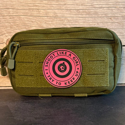 An olive green tactical pouch with a pink circular patch that reads 'I SHOOT LIKE A GIRL TRY TO KEEP UP' around a target graphic, set against a wood panel background.