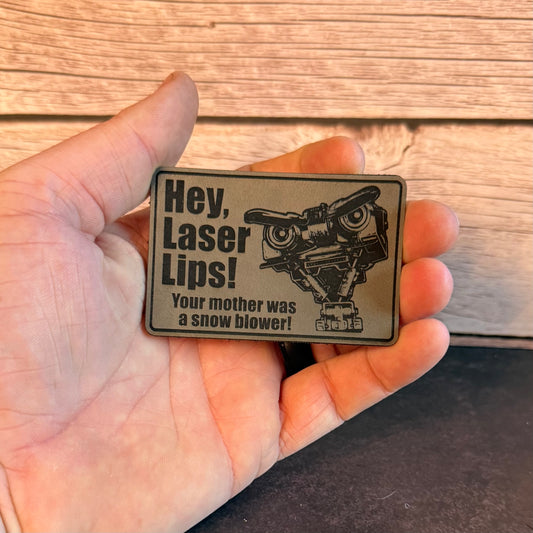 A hand holding a rectangular metal novelty buckle with a humorous message and a snow blower illustration, set against a wooden background.
