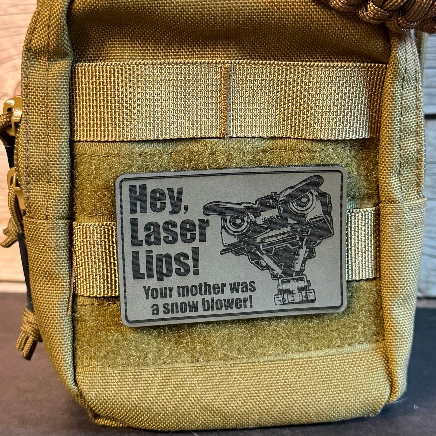 A close-up of a tan tactical pouch with a metal patch that reads 'Hey, Laser Lips! Your mother was a snow blower!' with a cartoon laser lip graphic. A woven strap is visible at the top, and the pouch is photographed against a wood plank background.