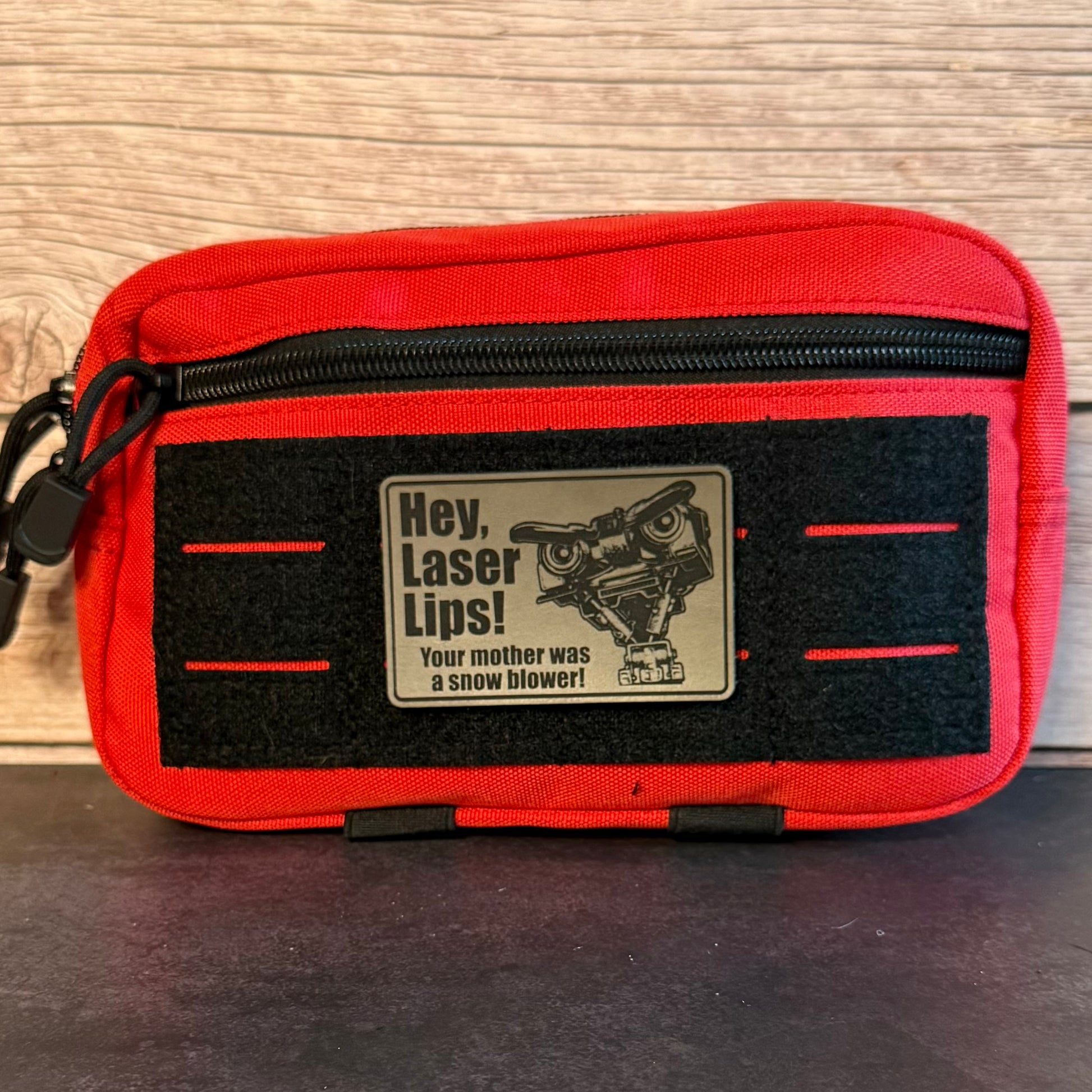 A red and black zippered pouch with a humorous patch that reads 'Hey, Laser Lips! Your mother was a snow blower!' featuring a cartoon snowmobile graphic, set against a wood panel background.