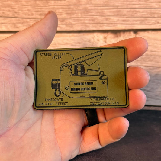 A hand holding a rectangular olive green metal tag with black printed diagrams and text reading 'STRESS RELIEF LEVER' and 'STRESS RELIEF FIRENG DEVICE MS7'.