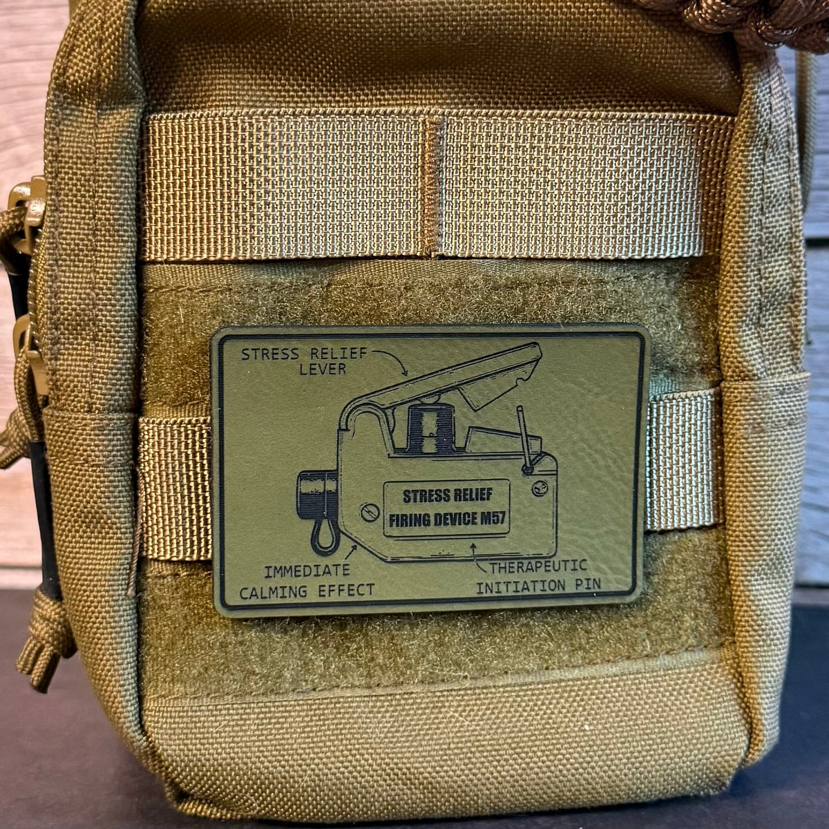 Close-up of a tan tactical pouch with a woven strap across the top and a rectangular patch on the front showing a stress relief lever diagram and the text 'STRESS RELIEF LEVER' and 'STRESS RELIEF THROUGH DEVICE NO. 1'.