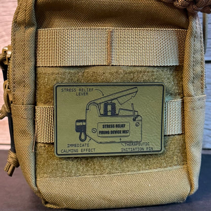 Close-up of a tan tactical pouch with a woven strap across the top and a rectangular patch on the front showing a stress relief lever diagram and the text 'STRESS RELIEF LEVER' and 'STRESS RELIEF THROUGH DEVICE NO. 1'.