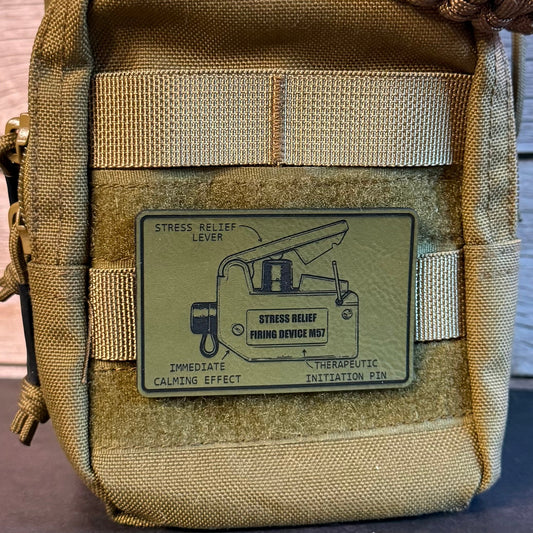Close-up of a tan tactical pouch with a woven strap across the top and a rectangular patch on the front showing a stress relief lever diagram and the text 'STRESS RELIEF LEVER' and 'STRESS RELIEF THROUGH DEVICE NO. 1'.
